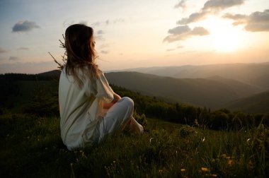 Kadın huzur içinde çimenli bir tepede oturur altın ışığın manzarayı yıkamasını seyreder. Yuvarlanan dağlar ve yemyeşil bir ağaç akşamları sakin bir atmosfer yaratır..