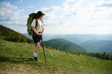 Bir kadın çimenli bir tepede büyük bir sırt çantası ve yürüyüş direkleri ile dikiliyor, uzak dağların manzarasının ve parlak bir gökyüzünün altındaki kır kulübesinin tadını çıkarıyor. Dışarıyı kucaklıyor..