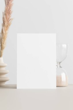 Üzerinde pampa süslemesi ve bej masa üzerinde kum saati olan beyaz davetiye maketi. 5x7 oranı, A6, A5 'e benziyor..