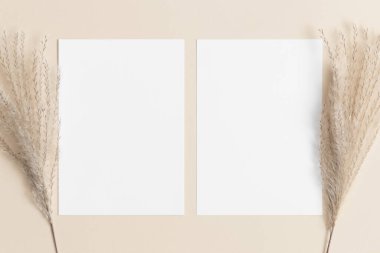 Bej masanın üzerinde sazlık pampa süslemesi olan iki beyaz davetiye kartı. 5x7 oranı, A6, A5 'e benziyor..