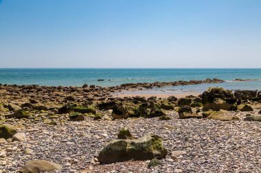 Dalgalar alçaldığında kayalık ve çakıl taşlı bir sahile tepeden bakan mavi gökyüzü
