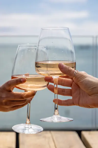Wine glasses raised in a toast at an outside bar, with the sea in the distance behind