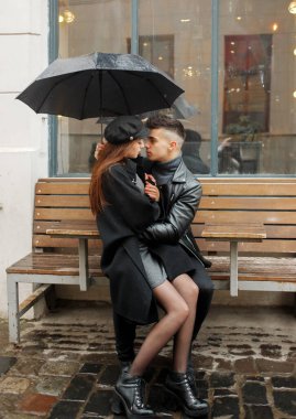 Young couple sitting near a cafe on a bench under an umbrella in rainy weather. Concept of love, romance and passion.