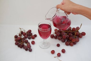 Kırmızı üzüm suyu beyaz arka planda taze üzümlü bir bardağa dökülüyor.