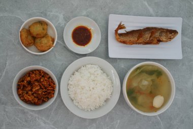 Bir tabak kızarmış balık, bir kase beyaz pirinç, köfteli bir kase sebze çorbası, küçük bir tabak kırmızı sos ve diğerleri, hepsi gri mermer bir yüzey üzerinde..