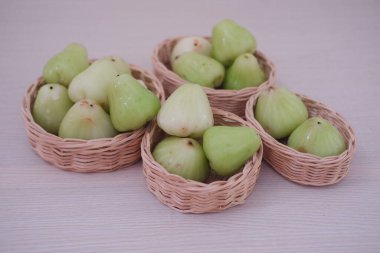 Açık yeşil su dolu küçük bir hasır sepet guava soluk ahşap bir yüzey üzerinde oturur. Meyvenin parlak, hafif engebeli bir dokusu ve konik bir şekli var..