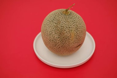 Koca bir kavun, koyu kırmızı arka planda, beyaz bir tabağa yerleştirilmiş.