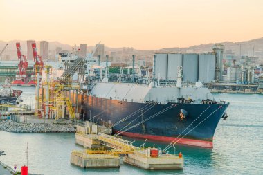 Büyük LNG (sıvılaştırılmış doğalgaz) tankeri, sarı boru hatları, depolama tankları ve vinçlerle bir endüstriyel liman terminaline demirlemiştir.