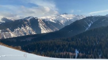 Aerial view of beautiful mountain ranges covered with snow and coniferous forests. Walk in the Karakol mountains. Blue sky and white clouds. Journey through amazing Kyrgyzstan.