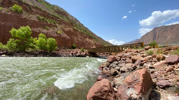 Fırtınalı bir dağ nehri köprünün altından akar. Büyük kayalar ve kıyıdaki ağaçlar. Güzel dağ vadisi. Dağların ve kanyonun manzarası. Kırgızistan 'ın inanılmaz doğası.