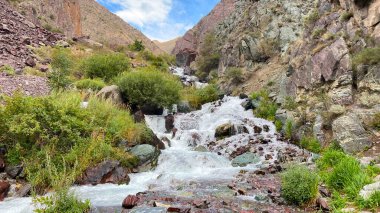Kırgızistan dağlarında küçük bir şelale. Güzel yaz manzarası. Kırgızistan 'daki kayaların, kayaların ve dağların manzarası. Mavi gökyüzü ve beyaz bulutlar.