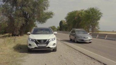 Yolun kenarında duran beyaz bir arabanın yakın çekimi, diğer arabaların koştuğu yerden geçiyor. Kırgızistan 'da arabayla seyahat ediyor. Yaz tatili.