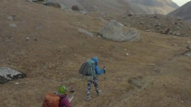Kamera kayalık bir dağ yolunda yürüyen iki turistin etrafında uçuyor. Bir kız ve büyük sırt çantalı ve sırıkla yürüyüş yapan bir adam yürüyüşe çıkıyorlar. Üst ve yan görünüm. Kırgızistan 'daki Ala-Archa Ulusal Parkı. Güzel çöl dağ manzarası.