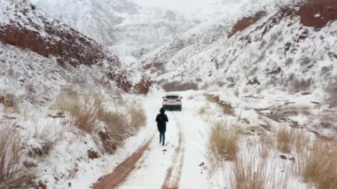 Siyah kapüşonlu bir kız, karlı bir dağ yolunda beyaz bir SUV 'ye doğru yürüyor ve etraftaki her şeyin fotoğrafını çekiyor. Konorchek Kanyonu. Kırgızistan 'a git. Güzel manzara.