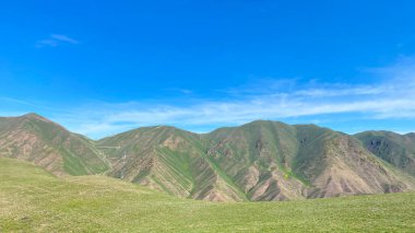 Beautiful views around the Konorchek canyons in Kyrgyzstan. The mountains are covered with green grass and rocks. The sky is clear and blue. The landscape is peaceful and serene, the mountains rise above the grassy plain clipart