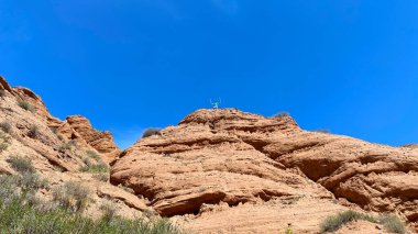 A man stands on top of a rocky hill, admiring the scenery. The red canyons of Konorchek. Travel to Kyrgyzstan. The sky is clear and blue, the sun is shining brightly. clipart