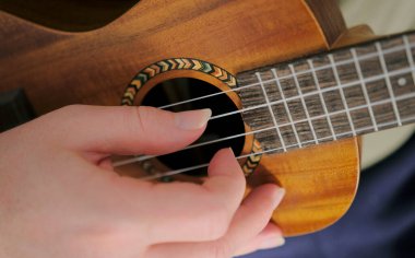 Ukulele 'nin boynundaki telleri koparan bir elin yakın çekimi. Adam ukulele çalıyor. Ukulele küçüktür ve kahverengi bir kapağı vardır. Müzisyenin eli bir ukulelenin boynunda.