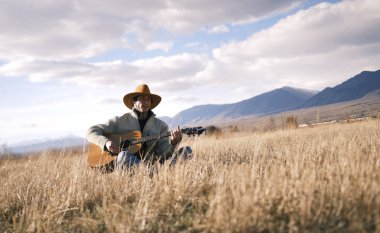 Deneyimli bir kovboy kuru otların arasında oturur ve gitar çalar. Kovboy şapkalı bekar bir adam elinde bir gitar tutuyor. Gökyüzü bulutlu, dağlar ve tepeler yükseliyor..