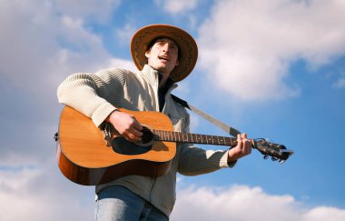 Genç bekar bir adam, mavi gökyüzü ve beyaz bulutların arkaplanına karşı gitarla şarkılar söylüyor, aşağıdan bir manzara. Özgürlük ve yaratıcılık ruhu. Gitarist parmaklarını tellerin üzerinde gezdirerek gitar çalar..