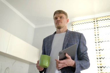 Ofiste çalışmak için acele eden bir iş adamı. Kendine güvenen, takım elbiseli, elinde tablet ve elinde yeşil kahve kupası olan başarılı bir adam, sabahın erken saatlerinde mutfakta duruyor. Bir profesyonel. Genç bir uzman.