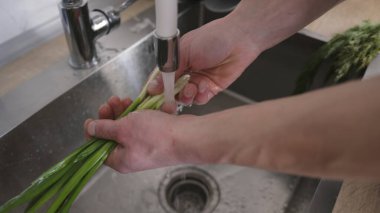 Suyun altında scallion yıkayan bir adamın yakın çekimi. Adam mutfağı işletiyor. Yemek pişiriyorum. Yeşillikleri yıkıyorum.