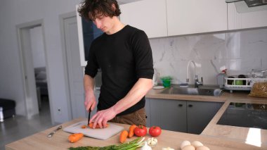 Genç ve yakışıklı bir öğrenci şık bir mutfakta kendi yemeğini hazırlar. Tahta bir tezgahta sebze doğruyor. Sebzeler arasında havuç, domates ve yeşil soğan yer alıyor..
