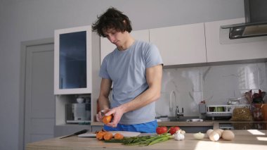 Beyaz bir adam sebze soyacağı ile havuç soyardı. Bir vejetaryen ahşap bir masada sebze keser. Mavi gömlek ve mavi şort giyiyor..