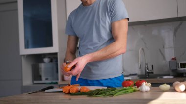 Şık bir mutfakta lezzetli bir yemek pişirmek. Tahta bir masada sebze doğrayan genç bir şefin yakın çekimi. Mavi gömlek ve mavi şort giyiyor..