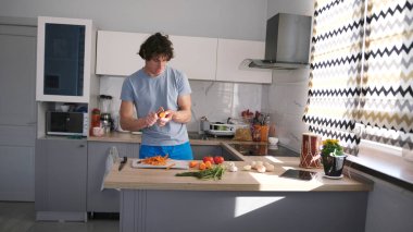 Neşeli bir adam mutfakta dans eder ve mutfak masasında sebze keser. Lezzetli bir sebze yemeği. Mutfakta mikrodalga fırın ve lavabo var. Adam mavi gömlek ve mavi şort giyiyor..