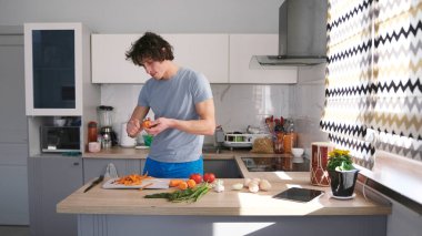Beyaz bir öğrenci şık, parlak bir mutfakta yemek pişirir. O, ahşap bir masada sebze keser. Mutfak mikrodalga fırın ve tost makinesi gibi cihazlarla donatılmıştır. Adam mavi bir gömlek giyiyor.