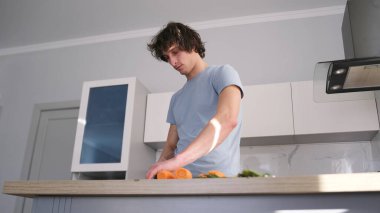 Aşağıdaki manzarada yakışıklı bir gay mutfakta taze havuç kesiyor. Mutfak şık, temiz ve bakımlı, tezgah ve dolaplarla dolu. Adam işine odaklanmış.