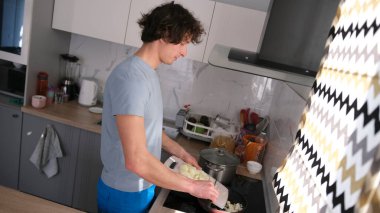 Çekici genç bir adam dilimlenmiş soğanları kızartma tavasına dolduruyor. Bir adam mutfakta soğan kızartıyor, ocakta bir tencere var. Mutfak iyi donatılmış. Adam mavi gömlek ve mavi pantolon giyiyor..