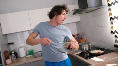 Mavi gömlekli yakışıklı bir adam mutfakta yemek pişirir, elinde tava kapağıyla neşeyle dans eder. Neşeli bir bekar. Öğrenci yemek hazırlar.