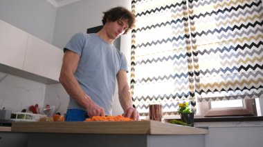Mutfaktaki sevimli eşcinsel adam tahta kesme tahtasıyla havuç kesiyor. Lezzetli bir öğle yemeği pişiriyorum. Mutfak modern bir şekilde ışıklandırılmış ve dekore edilmiş..