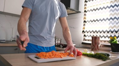 Kesme tahtasında havuç kesen bir adamın yakın çekimi. Aşçı akşam yemeği hazırlıyor. Mutfakta bir öğrenci var. Adam mavi gömlek ve mavi şort giyiyor..