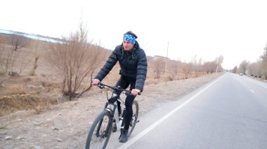 Mutlu bisikletçi eğitime gitti. Kasvetli bir kırsal bölgenin arka planında bisiklete binen bir adam. Kafasında mavi bir bandana var..