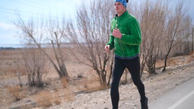 Yakışıklı bir adam yeşil bir ceket ve kafasında mavi bir bandana ile bir tarlada koşuyor. Açık hava egzersizi. Sportif bir yaşam tarzı. Kırsal bölgelerde atölye çalışması