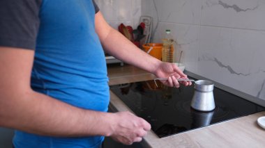 Adamın biri kokulu kahve yapmak için indüksiyon hobisine Türk koyuyor. Sabah rutinleri. Mavi bir gömlek giyiyor ve küçük bir göbeği var. Erkek kahvaltısı.