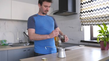 Mavi gömlekli genç bir adam cam kavanozdan bir kaşık kahve alıp bir fincana döker ve kahve yapar. Mutfak temiz ve bakımlı. Masanın üzerinde saksı var..