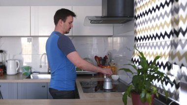 Yakışıklı bir eşcinsel mutfakta durur ve kahve yapar. Mutfakta soba, lavabo ve saksı bitkisi var. Adam mavi bir gömlek giyiyor ve işine odaklanmış durumda..