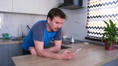 Beyaz bir adam mutfak masasında duruyor ve cep telefonuna bakıyor. Mavi bir gömlek giymiş ve düzgün bir sakal ve bıyık takıyor. Kahve bekliyorum.