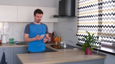 Yalnız beyaz bir adam elinde telefonla mutfakta duruyor ve bir Türk 'ün içinde kahve yapıyor. Mutfak temiz ve iyi döşenmiş, ve masanın üzerinde saksı bitkisi var..