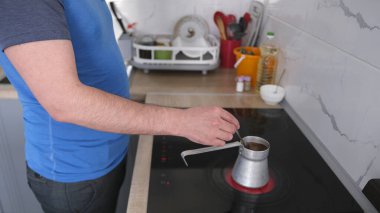 Bir adam mutfaktaki bir Türk 'te kahve yapıyor. Kahveyi karıştırmak için metal bir kahve demliği ve kaşık kullanıyor. Mutfak, lavabo ve buzdolabı da dahil olmak üzere çeşitli mutfak aletleriyle dolu..