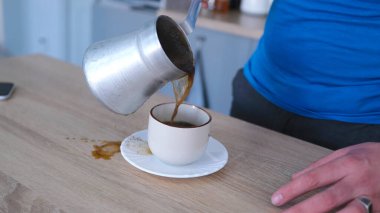 Güzel kokulu bir sabah. Bekar bir adamın kahvaltısı. Türk fincanından kahve döken bir öğrencinin ahşap bir masada çay tabağının üzerinde duran beyaz bardağa yakın plan fotoğrafı..