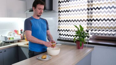 Sevimli bir adam mutfakta sebze soyucusuyla çiğ patates soyuyordu. Mavi bir gömlek giyiyor ve kendine akşam yemeği pişiriyor. Mutfakta lavabo, saksı ve pencere var..