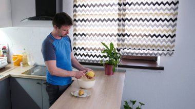 Bir öğrenci ahşap mutfak masasında çiğ patates soyuyor. Mutfak temiz ve bakımlı, mutfak masasında lavabonun yanında saksı bitkisi var. Adam mavi bir gömlek giyiyor.