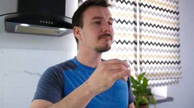 Mavi gömlekli, üzgün, uykulu bir adam mutfakta duran küçük bir bardaktan sıcak, kokulu kahve içer. Rahatlama ve zevk kavramı. Bir erkek rahat bir ortamda içki içmekten hoşlandığında.