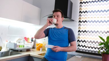 Mutfakta güzel kokulu bir sabah kahvesi içen mutlu beyaz bir adam. Mutfak temiz ve derli toplu. Adam mavi bir gömlek giyiyor ve elinde beyaz bir tabak tutuyor..