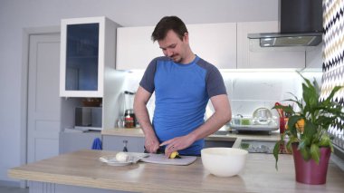 Gülümseyen genç bir adam mutfaktaki ahşap tezgahta çiğ patatesleri dilimlere ayırıyor. Mutfak iyi aydınlatılmış ve mutfak masasında saksı bitkisi var..