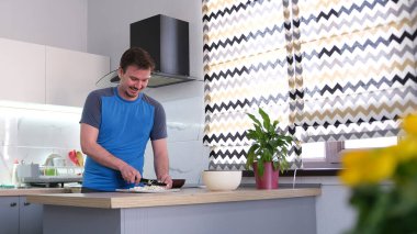 Gülen genç bir öğrenci öğle yemeği hazırlar. Bir adam şık bir mutfakta duruyor ve kesme tahtasında sebzeleri dilimliyor. Mutfak temiz ve bakımlı. Masanın üzerinde saksı var..